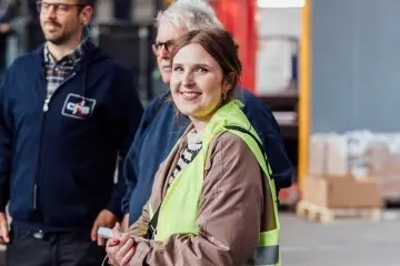 Eine weibliche Person, die in einer Halle steht und etwas in der Hand hält. Sie trägt eine Warnweste und lächelt dabei in die Kamera, Imke Thomann.