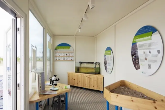 Die Innenseite eines Informationencontainers der CHS Container Group. Eine Seite des Containers ist komplett verglast. Es befinden sich verschiedene Exponate im Container und Schilder. 