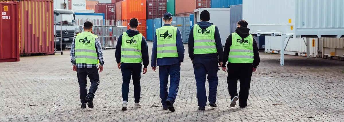 Fünf Personen laufen von der Kamera weg auf ein Containerdepot. Sie tragen alle Warnwestern auf denen hinten das Logo der CHS Container Group zu sehen ist.