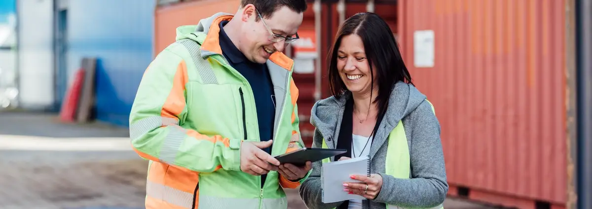 Zwei Personen stehen auf einem Containerdepot und tragen Warnwesten. Sie schauen beide auf ein Tablet und lachen dabei.