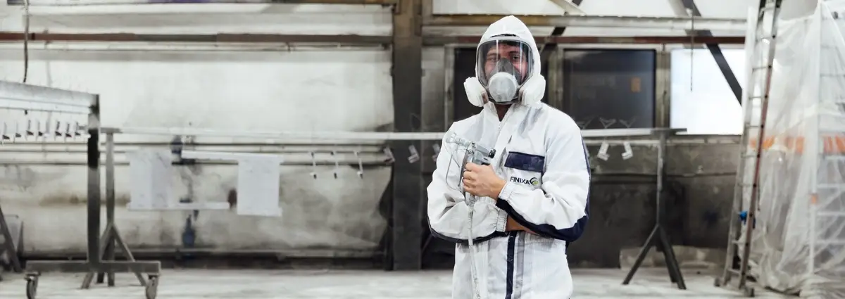 Eine männliche Person trägt eine Ausrüstung bzw. Schutzkleidung zum Lackieren und hat eine Farbpistole in der Hand. Er schaut frontal in die Kamera.