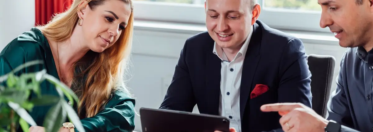 Drei Personen schauen gemeinsam auf ein Tablet. Sie befinden sich in einem Meeting und vor ihnen steht Kaffee und Kekse auf dem Tisch. 