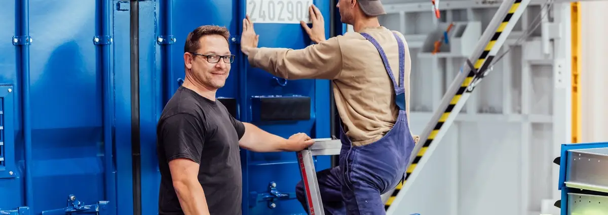 Zwei männliche Personen stehen an einem Container. Eine Person bringt gerade eine Nummer am Container an und steht mit dem Rücken zur Kamera. Die andere Person schaut in die Kamera und lächelt.