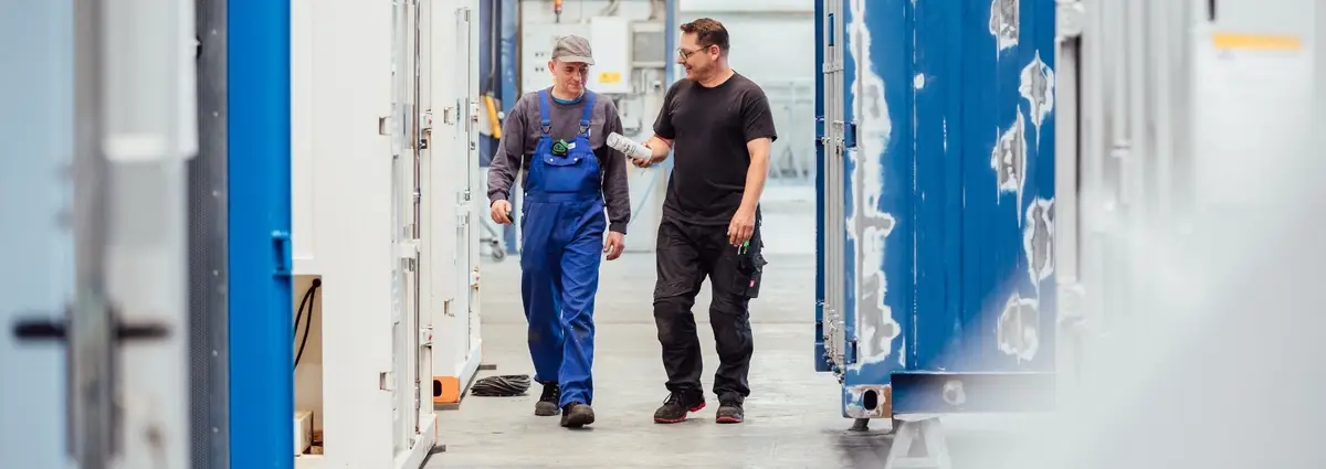 Zwei männliche Personen laufen zwischen Containern und unterhalten sich. Beide lächeln.