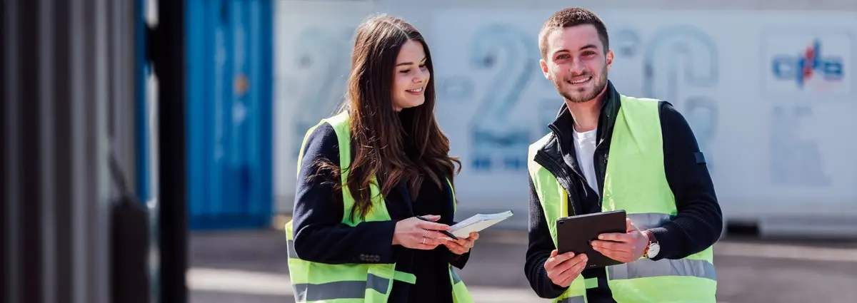 Eine weibliche und eine männliche Person. Die weibliche Person schaut auf das Tablet in der Hand der männlichen Person. Die männliche Person schaut in die Kamera. Beide tragen Warnwesten und lächeln.