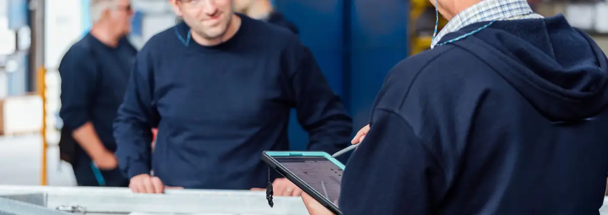 Zwei Männer lächeln sich an und stehen vor einem Stück Metall. Der eine Mann hält dabei ein Tablet in der Hand. 