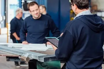 Zwei Männer lächeln sich an und stehen vor einem Stück Metall. Der eine Mann hält dabei ein Tablet in der Hand.