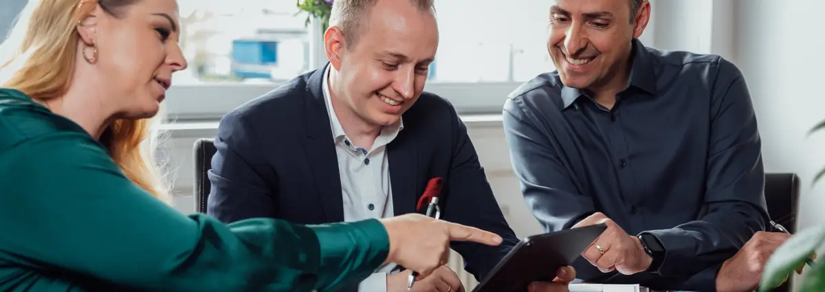 Drei Personen schauen gemeinsam auf ein Tablet. Sie befinden sich in einem Meeting und vor ihnen steht Kaffee und Kekse auf dem Tisch. 