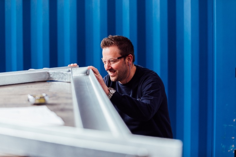 Eine Person, die mit einem Auge geschlossen über die Ecke eines metallischen Bauteils schaut und eine Schutzbrille trägt.