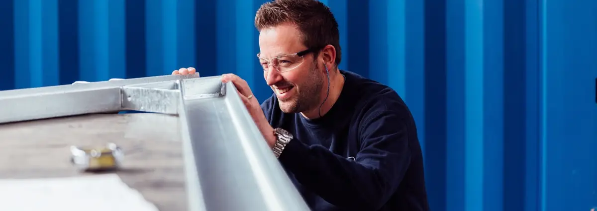 Eine Person, die mit einem Auge geschlossen über die Ecke eines metallischen Bauteils schaut und eine Schutzbrille trägt. 