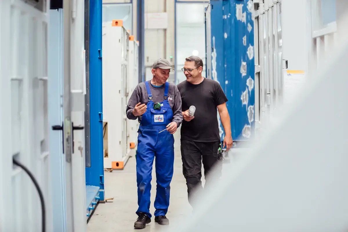 Zwei Mitarbeiter der CHS Container Handel GmbH unterhalten sich, während sie durch ein Lager mit Seecontainern gehen.