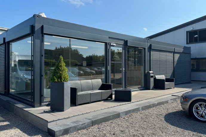 Büro (3) Ein Bürocontainer in einem dunkelgrau, der an einer Seite komplett verglast wurde. Vor ihm stehen Terrassenmöbel.