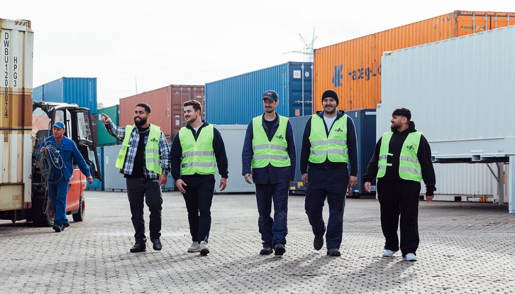 Fünf Personen tragen Warnwesten und laufen auf dem Depot der CHS Container Group auf die Kamera zu. Eine Person zeigt dabei auf einen Container.