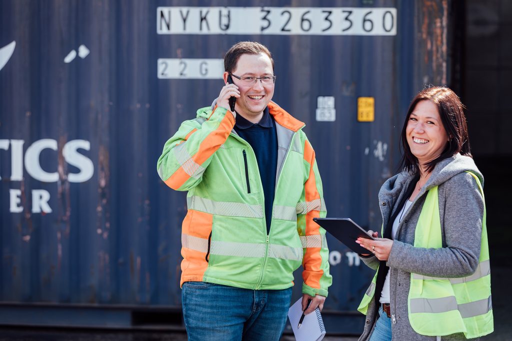 Zwei Personen stehen vor einem Container in Warnwesten und lächeln in die Kamera. Eine Person telefoniert und die andere hat ein Tablet in der Hand.