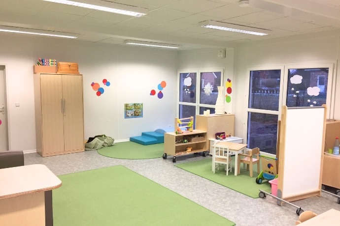 Kindergärten (2) Die Innenansicht einer Schule gebaut aus Containern der CHS Container Group. Man sieht mehrere Teppiche, Spielzeuge und Zeichnungen an den Fenstern und Wänden.