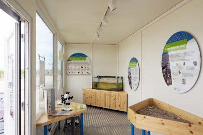 Projekt Sanitär- und Informationscontainer (2) Die Innenseite eines Informationencontainers der CHS Container Group. Eine Seite des Containers ist komplett verglast. Es befinden sich verschiedene Exponate im Container und Schilder.