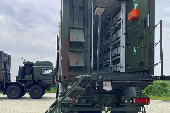Ein Werkstattcontainer für die militärische Anwendung der CHS Container Group. Der Container hat ein Tarnmuster und ist hinten geöffnet. Eine Treppe hinten und in der Mitte des Containers führt nach oben in den Container.