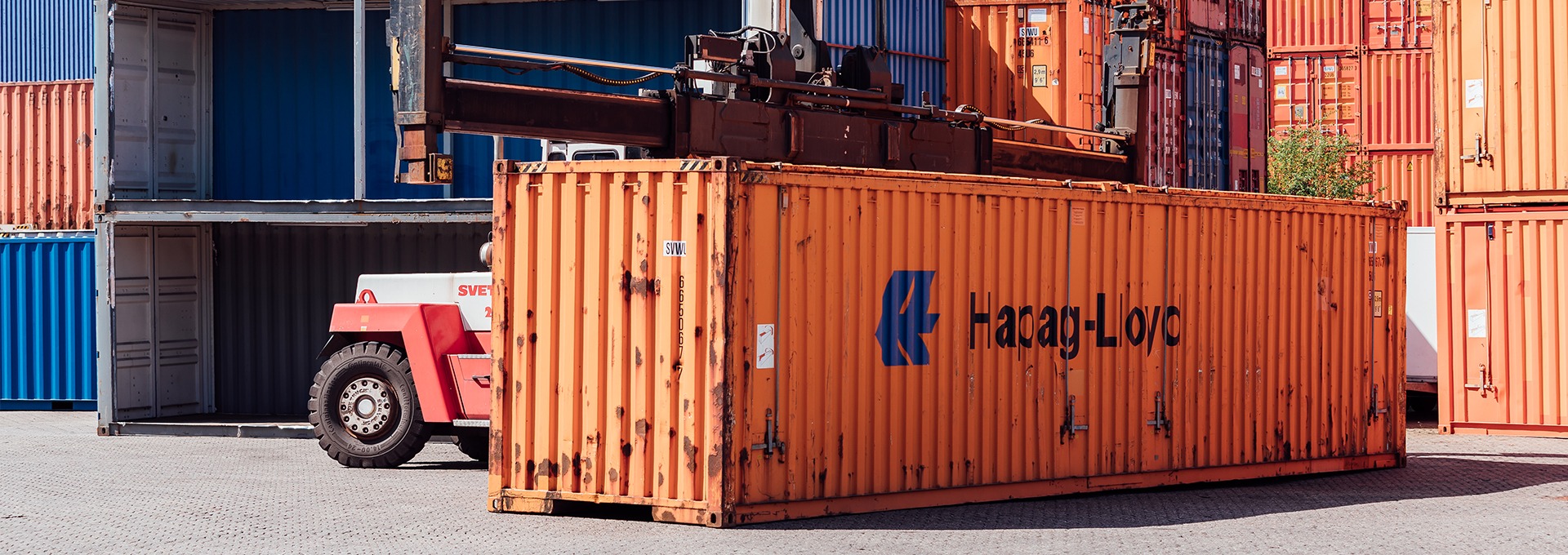 Ein orangener Container steht auf dem Containerdepot der CHS Container Group und wird von einem Gabelstapler hochgenommen.