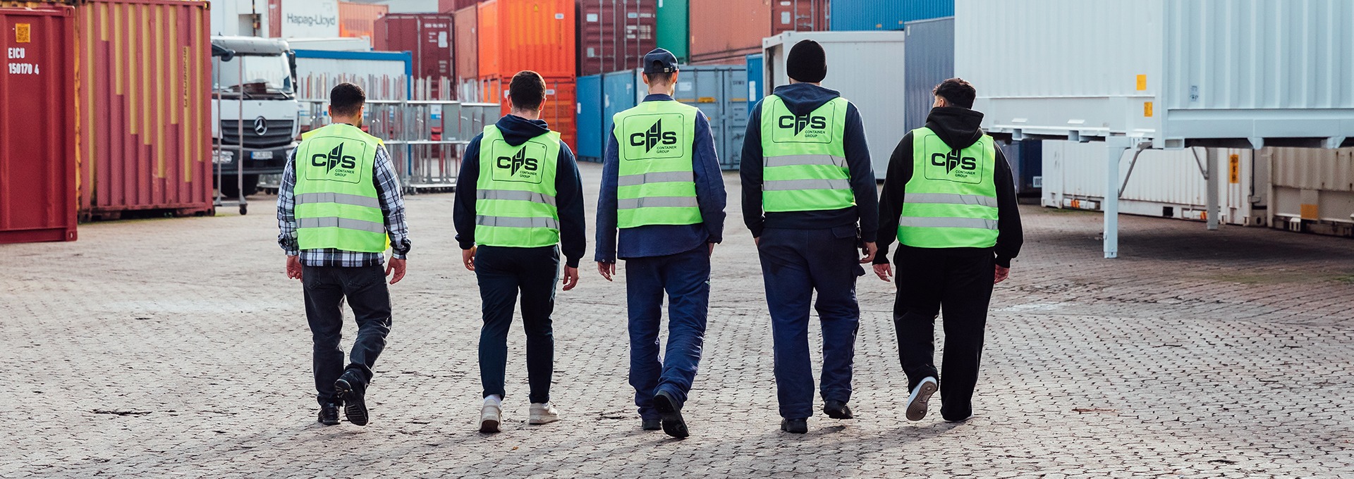 Fünf Personen laufen von der Kamera weg auf ein Containerdepot. Sie tragen alle Warnwestern auf denen hinten das Logo der CHS Container Group zu sehen ist.