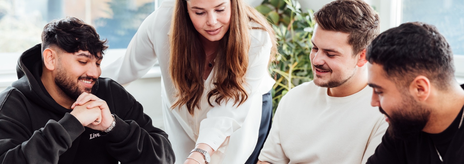 Drei männliche und eine weibliche Person. Die Weibliche Person zeigt etwas auf einem Blatt auf dem Tisch. Die drei männlichen Personen schauen auf das Blatt. Alle lächeln.