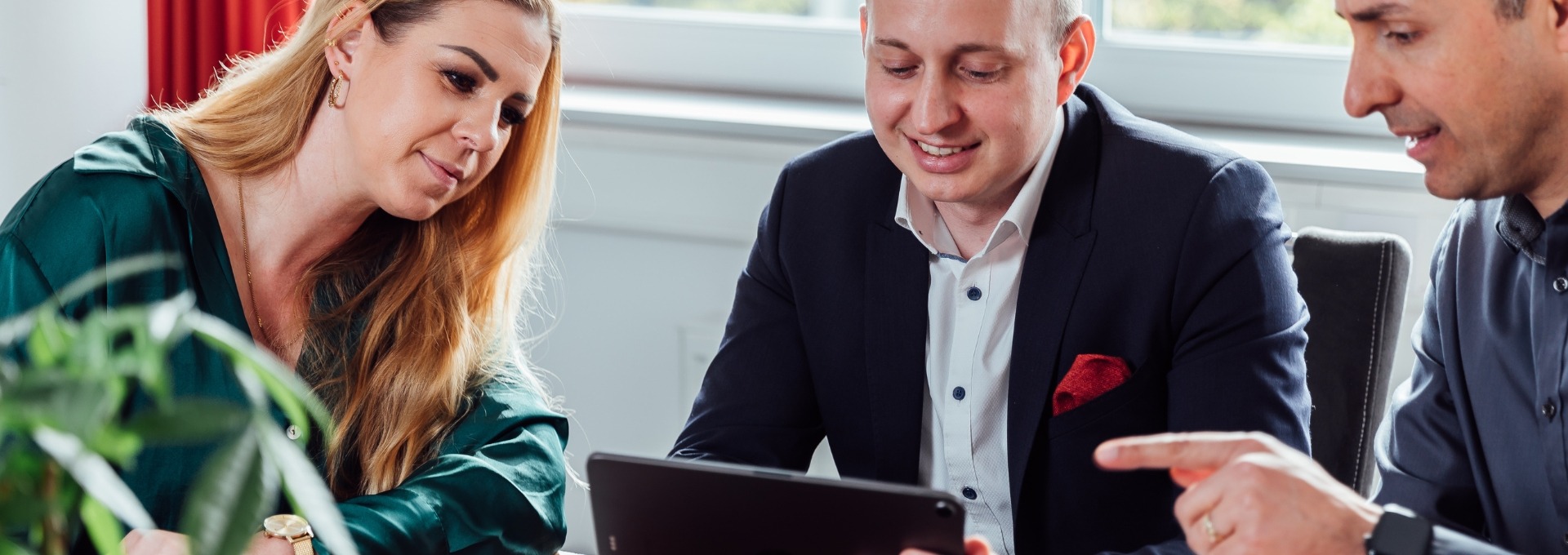 Drei Personen schauen gemeinsam auf ein Tablet. Sie befinden sich in einem Meeting und vor ihnen steht Kaffee und Kekse auf dem Tisch. 