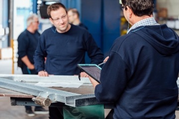 Zwei Männer lächeln sich an und stehen vor einem Stück Metall. Der eine Mann hält dabei ein Tablet in der Hand.