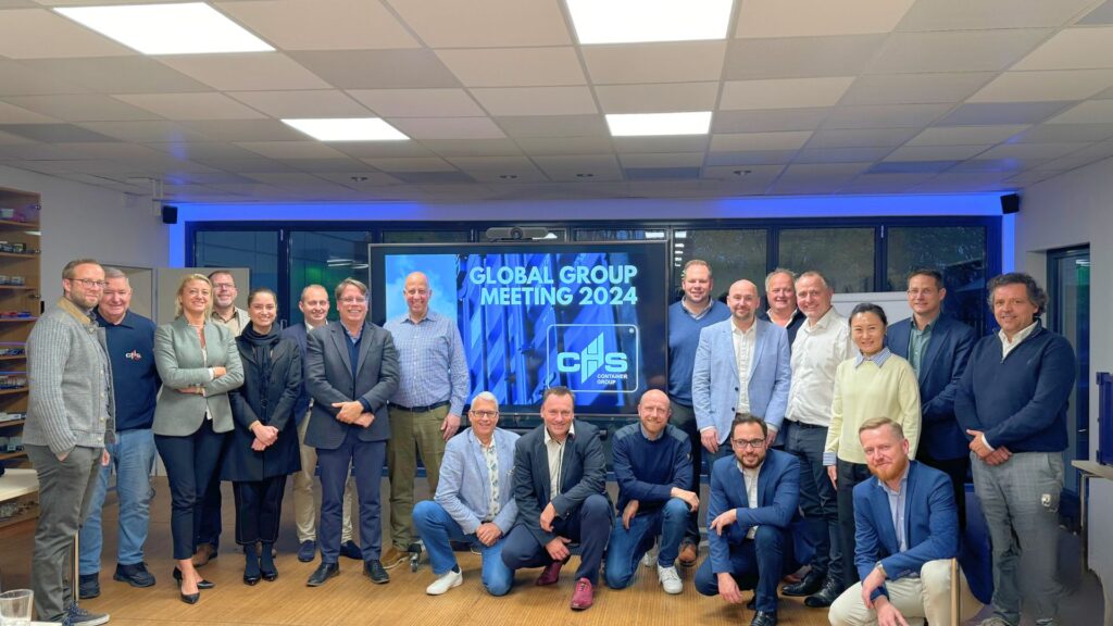Teilnehmer des Global Group Meetings 2024 stehen im Veranstaltungszentrum vor einem Bildschirm. Auf ihm steht "Global Group Meeting 2024"
