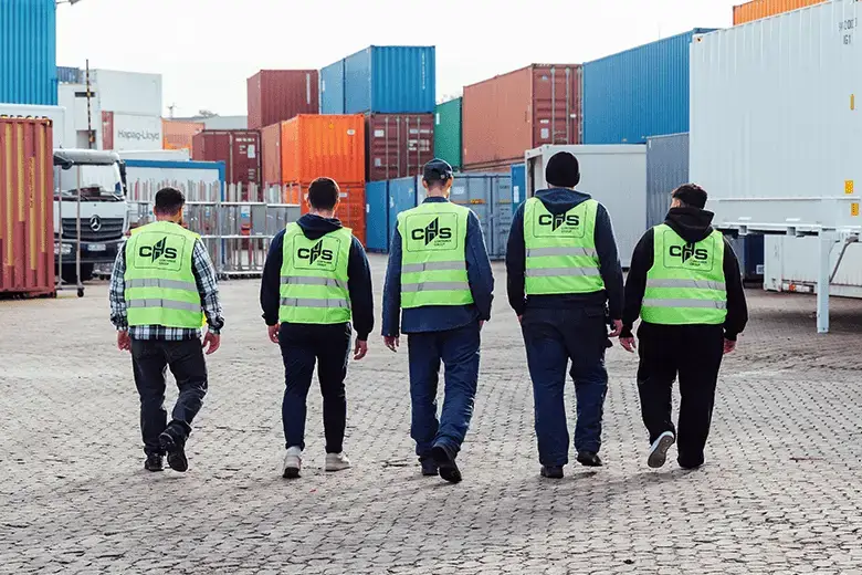 Fünf Personen laufen von der Kamera weg auf ein Containerdepot. Sie tragen alle Warnwestern auf denen hinten das Logo der CHS Container Group zu sehen ist. 