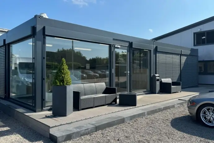 Ein Bürocontainer in einem dunkelgrau, der an einer Seite komplett verglast wurde. Vor ihm stehen Terrassenmöbel. 