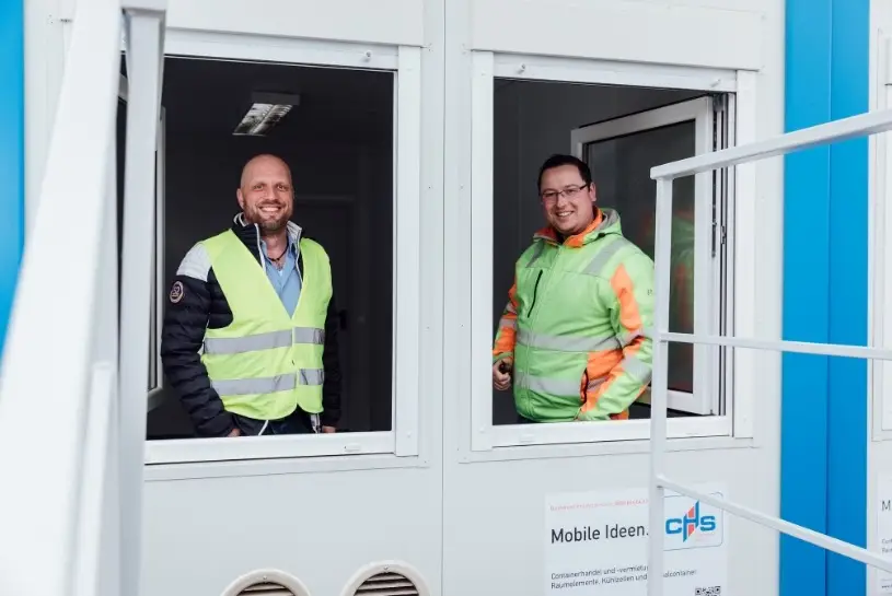 Zwei Männern stehen in Warnwesten in einem Container und schauen aus dem geöffneten Fenster in die Kamera und lächeln dabei. 