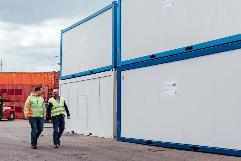 Zwei Männer laufen in Warnwesten vor Bürcontainern der CHS Container Group entlang und lächeln sich dabei an. 