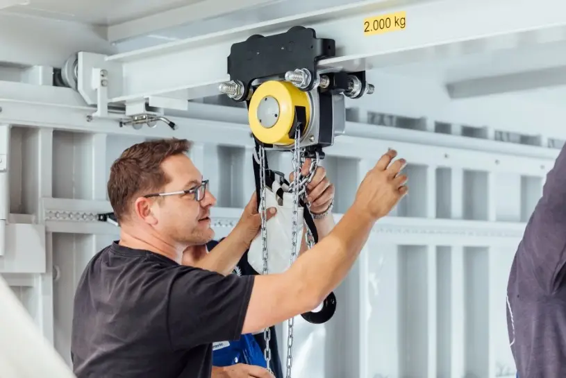 Ein Mann steht in einem Container und hat eine Hand an einer Winde von der eine Eisenkette abgeht. Er zeigt mit einer Hand zur Decke des Containers