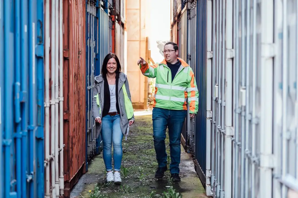 Zwei Personen in Warnweste laufen zwischen Containern entlang. Eine Person lächelt in die Kamera und die andere zeigt auf einen Container.