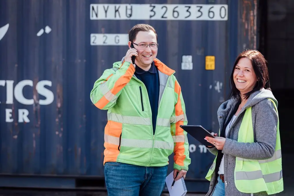 Zwei Personen stehen vor einem Container in Warnwesten und lächeln in die Kamera. Eine Person telefoniert und die andere hat ein Tablet in der Hand.