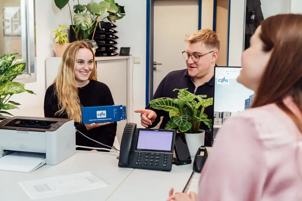 Zwei Personen sitzen an einem Tisch. Eine Person zeigt einen Miniaturcontainer der CHS Container Group der Person, der sie gegenübersitzt. Die andere Person zeigt auf den Container.