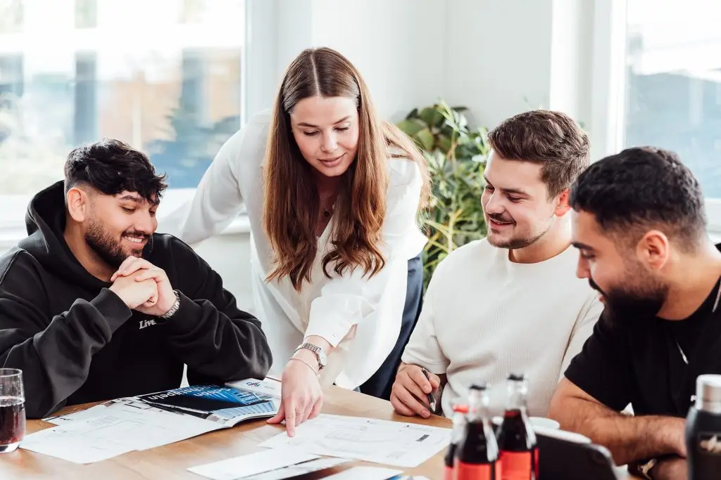 Drei männliche und eine weibliche Person. Die Weibliche Person zeigt etwas auf einem Blatt auf dem Tisch. Die drei männlichen Personen schauen auf das Blatt. Alle lächeln.