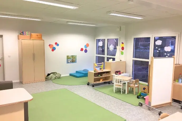 Die Innenansicht einer Schule gebaut aus Containern der CHS Container Group. Man sieht mehrere Teppiche, Spielzeuge und Zeichnungen an den Fenstern und Wänden.