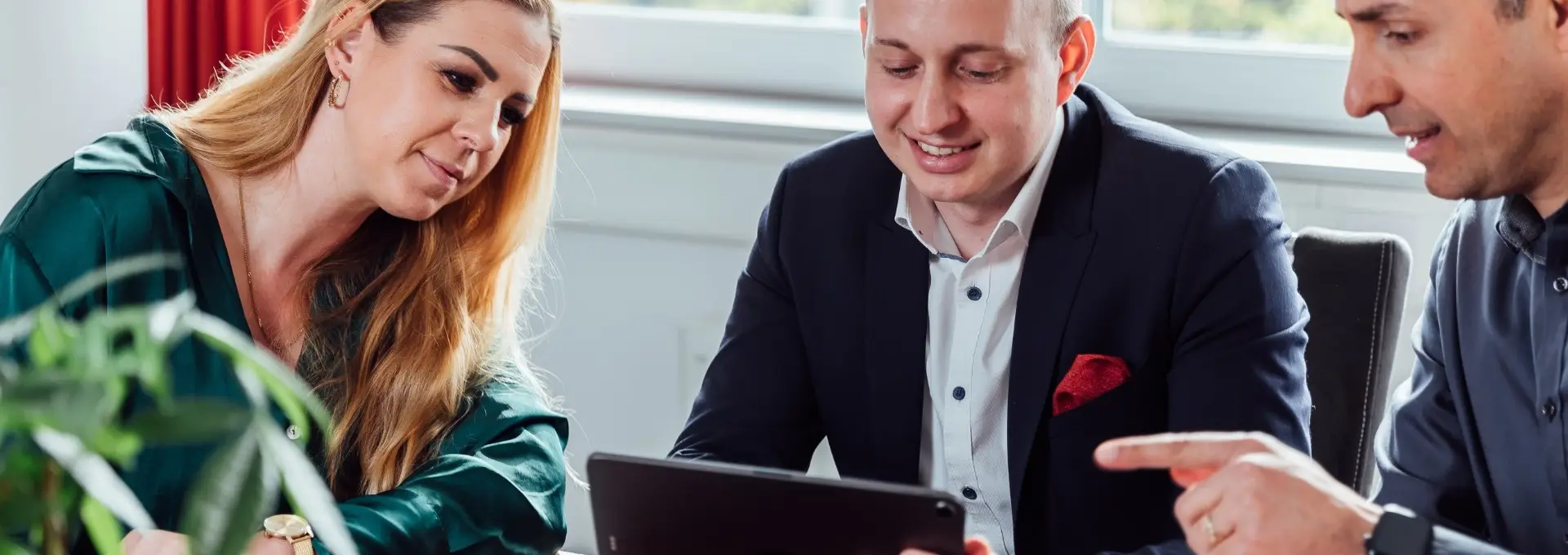 Compliance Header Desktop Drei Personen schauen gemeinsam auf ein Tablet. Sie befinden sich in einem Meeting und vor ihnen steht Kaffee und Kekse auf dem Tisch.