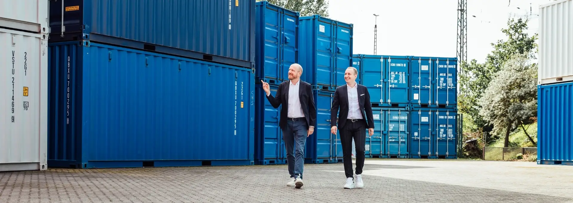 Zwei Personen laufen zwischen Containern und eine Person zeigt auf die Container. Beide lächeln.