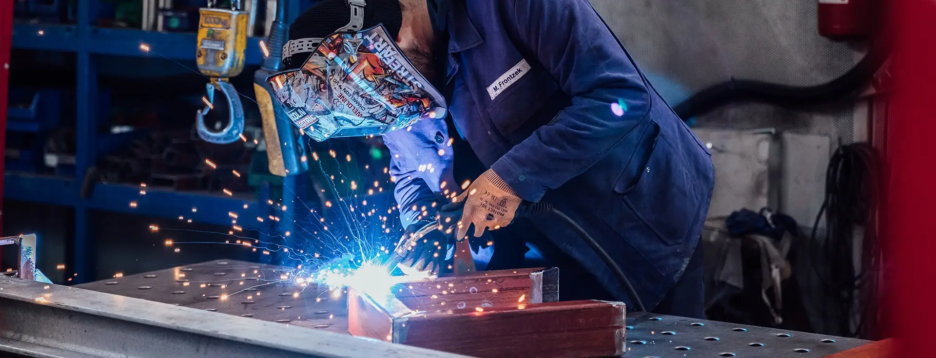 Ein Schweißer trägt eine Schweißermaske und schweißt ein Stück Metall von dem Funken ausgehen.